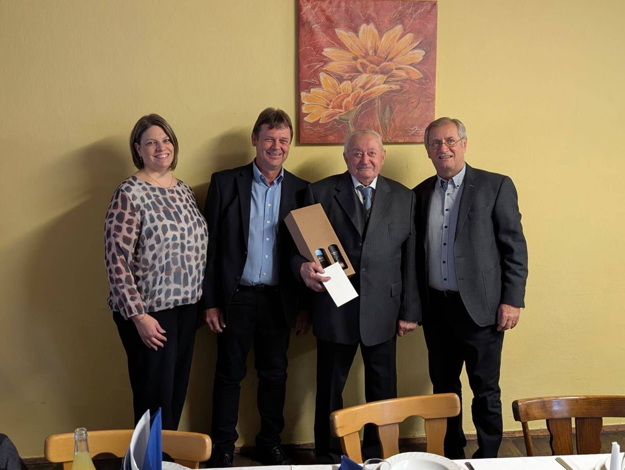 Vier Personen stehen gemeinsam in einem Raum und lächeln für ein Foto. Sie sind alle feierlich gekleidet. Ein Mann auf der rechten Seite hält eine Schachtel in der Hand. Hinter ihnen befindet sich eine gelbe Wand mit einem Blumenbild.