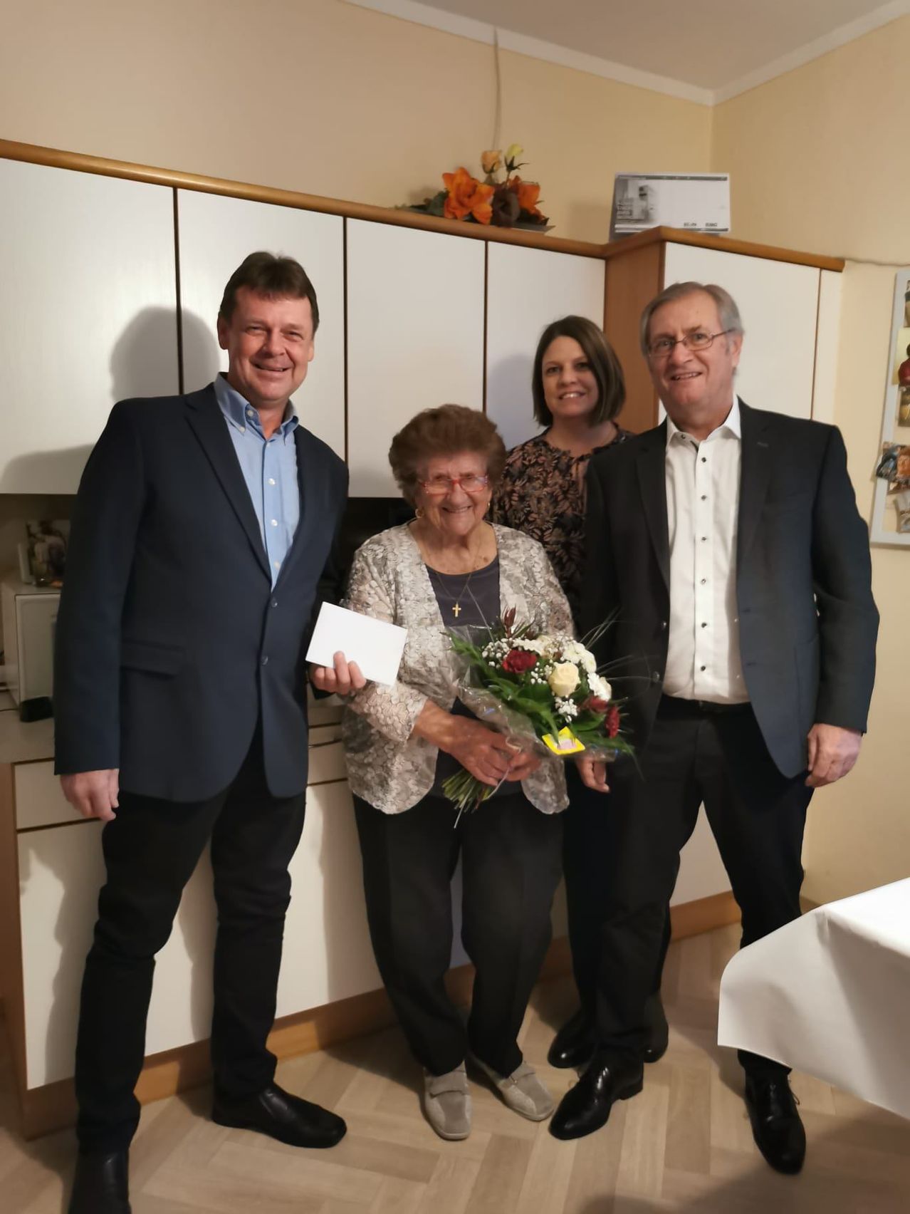 Vier Menschen stehen lächelnd in einem Raum. Eine Frau in der Mitte hält einen Blumenstrauß und ein Stück Papier. Der Mann auf der linken Seite hält ein Stück Papier. Hinter ihnen sind weiße Schränke und eine weiße Tischdecke zu sehen.