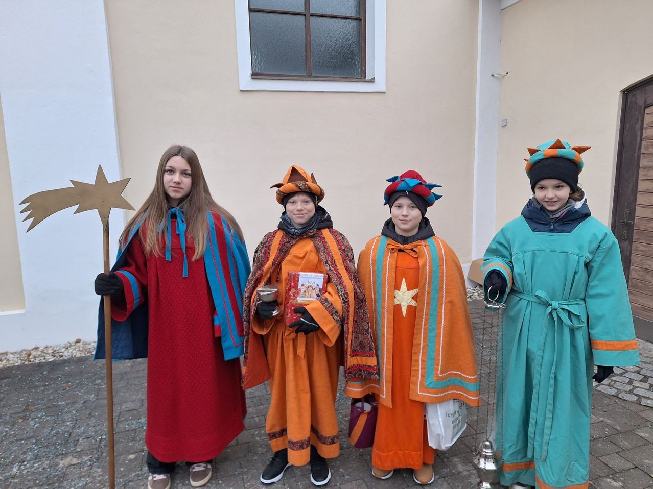 Vier Kinder in Kostümen stehen vor einem Gebäude, möglicherweise für eine Aufführung. Sie halten Requisiten, und eines hält ein Buch. Das Mädchen auf der linken Seite hält einen Stern.