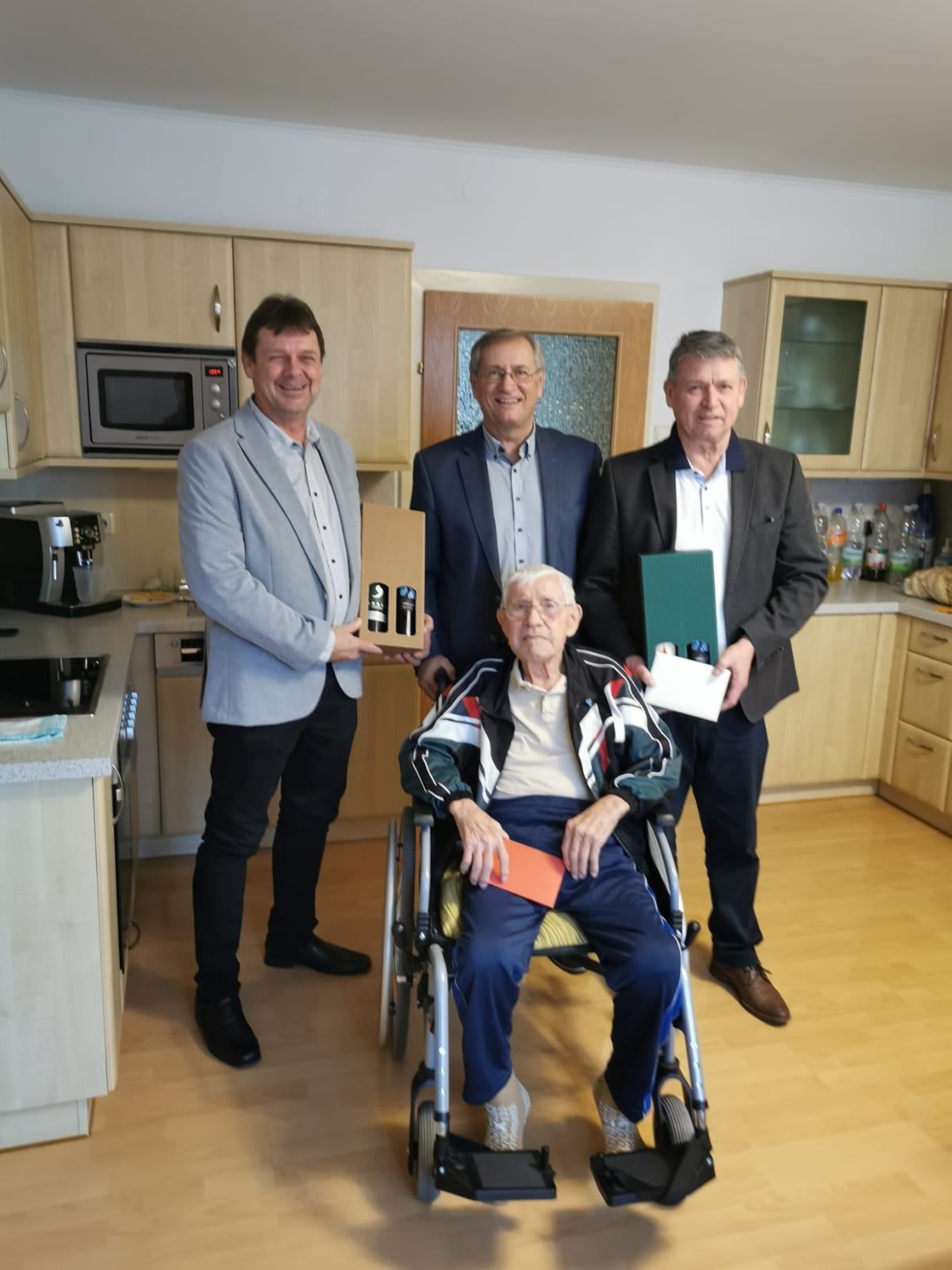 Vier Männer in einer Küche, einer im Rollstuhl, alle lächeln. Drei halten eine Box, ein Buch und einen Beutel.