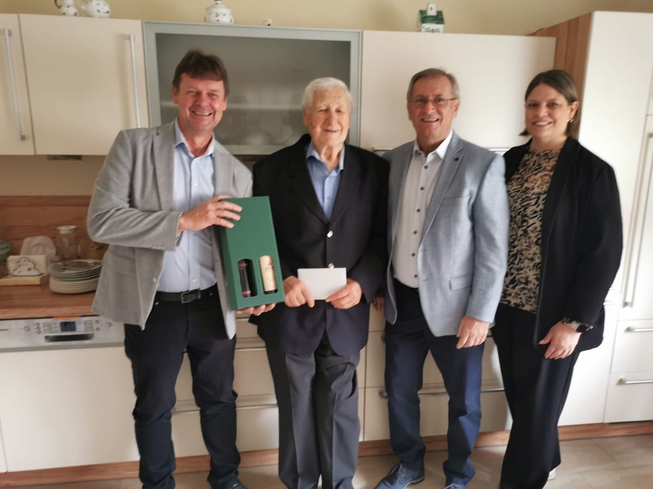 Vier Menschen stehen in einer Küche. Der Mann in der Mitte hält eine grüne Box mit zwei Flaschen darin. Der Mann auf der rechten Seite lächelt und hält ein weißes Papier.