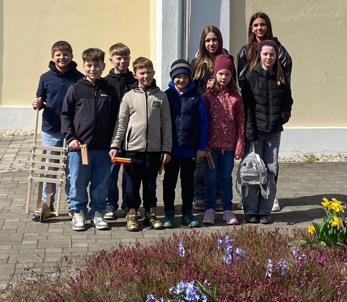 Eine Gruppe von Kindern posiert für ein Foto draußen, mit Holzstäben in der Hand. Sie stehen vor einem Gebäude mit einem Blumenbeet im Vordergrund.