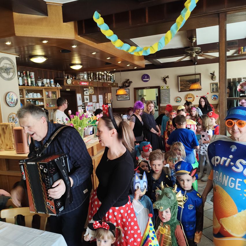 In einem lebhaften Restaurant spielt ein Mann Akkordeon, während eine Frau lächelt. Eine Gruppe von Kindern in Kostümen versammelt sich um sie herum. Bunte Luftballons hängen von der Decke, und Regale mit verschiedenen Gegenständen sind zu sehen.