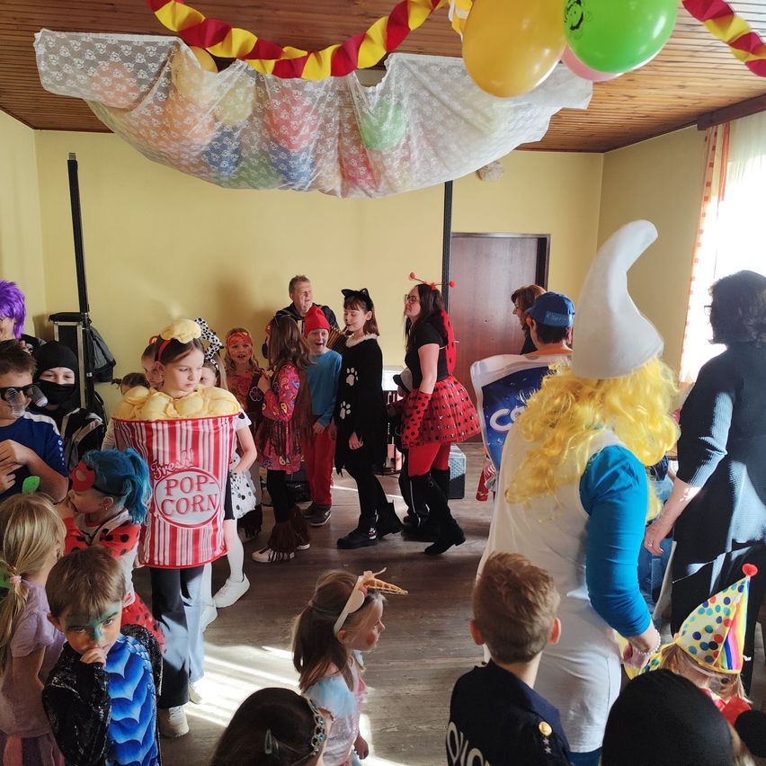 Eine Gruppe von Kindern in Kostümen und mit Schminkgesichtern, einige mit Masken, versammeln sich in einem Raum mit einem Popcorn-Maskottchen. Bunte Luftballons hängen von der Decke.