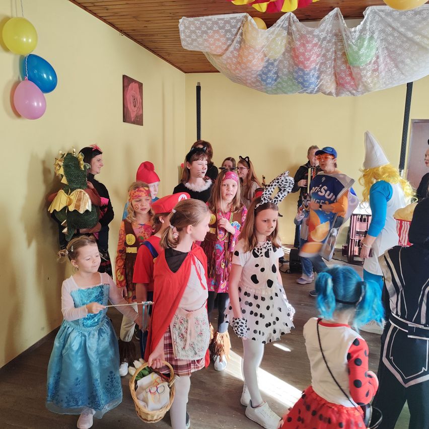 Eine Gruppe von Kindern in verschiedenen Kostümen steht in einem Raum mit gelben Wänden, Ballons und Dekorationen. Sie scheinen auf einer Kostümparty oder einem Event zu sein.