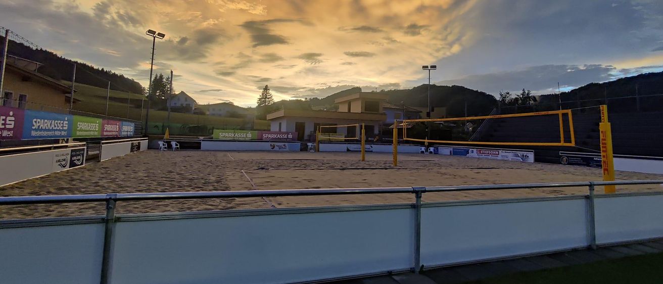 Ein leerer Beachvolleyballplatz im Freien mit einem Sonnenuntergang im Hintergrund. Der Himmel ist orange und bewölkt. Es gibt zwei Stangen mit Netzen.