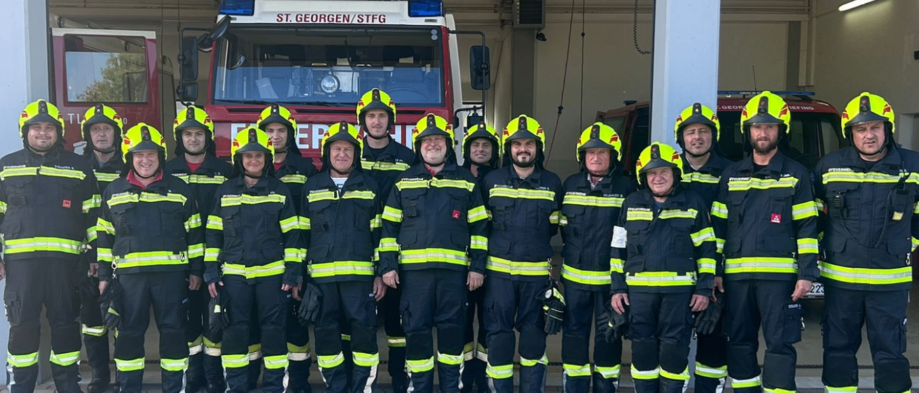 Eine Gruppe Feuerwehrleute in Uniform posiert für ein Foto vor einem Feuerwehrwagen. Der Wagen trägt den Namen St. Georg/STFG.