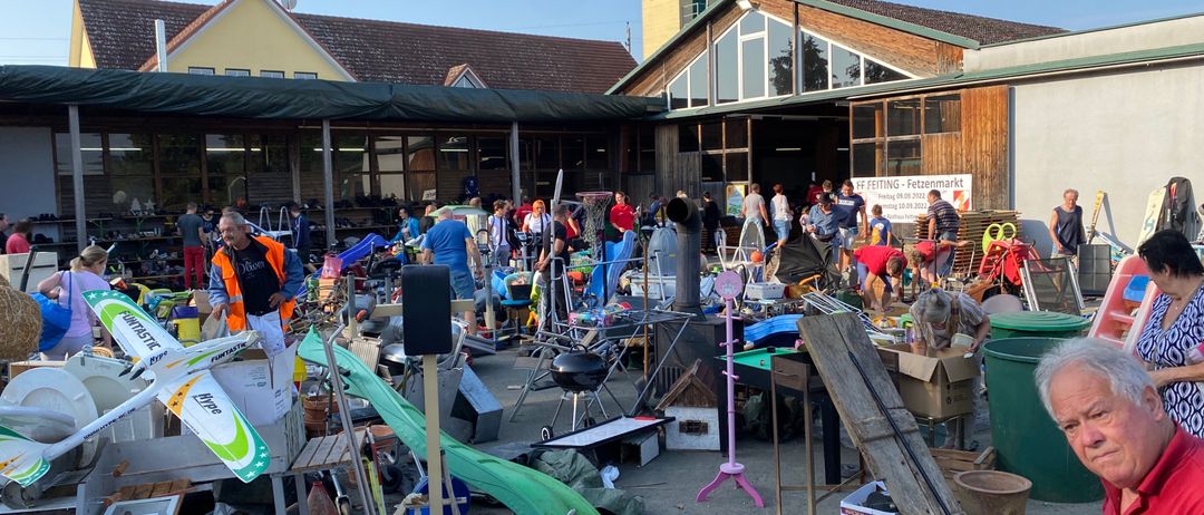 Ein Flohmarkt findet vor einem Gebäude statt. Es sind verschiedene Gegenstände ausgestellt, darunter Spielzeug, Kisten und andere Objekte. Viele Menschen sind anwesend, einige kaufen und andere verkaufen. Der Himmel ist klar.