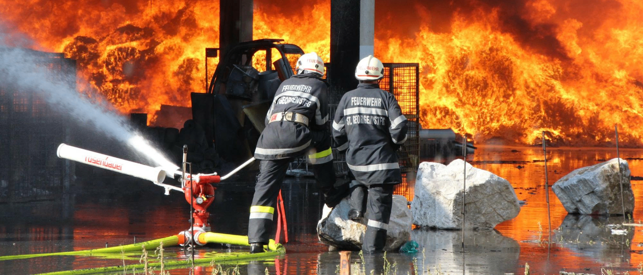 Zwei Feuerwehrleute bekämpfen ein großes Feuer mit Wasserwerfern. Das Feuer ist so intensiv, dass es ein nahes Fahrzeug zerstört hat. Sie tragen Schutzuniformen und Helme.