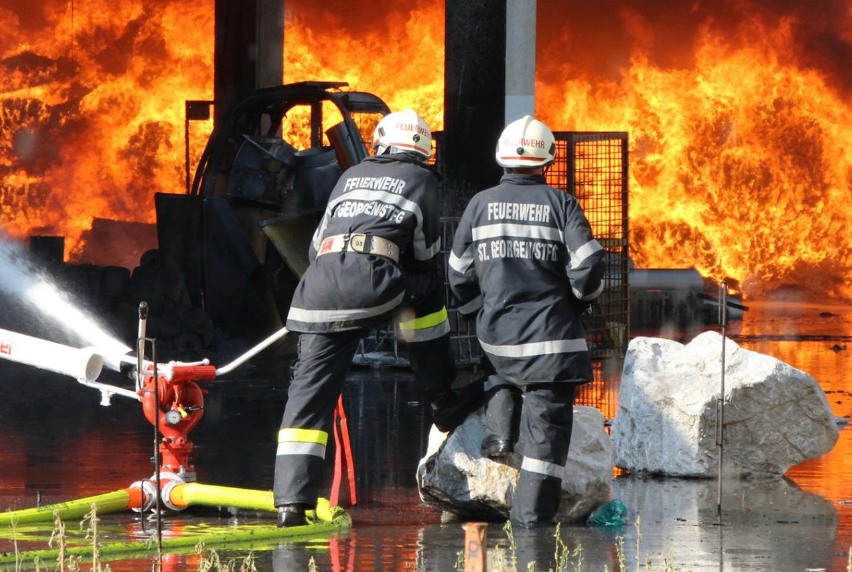 Zwei Feuerwehrleute bekämpfen ein großes Feuer mit Wasserwerfern. Das Feuer ist so intensiv, dass es ein nahes Fahrzeug zerstört hat. Sie tragen Schutzuniformen und Helme.