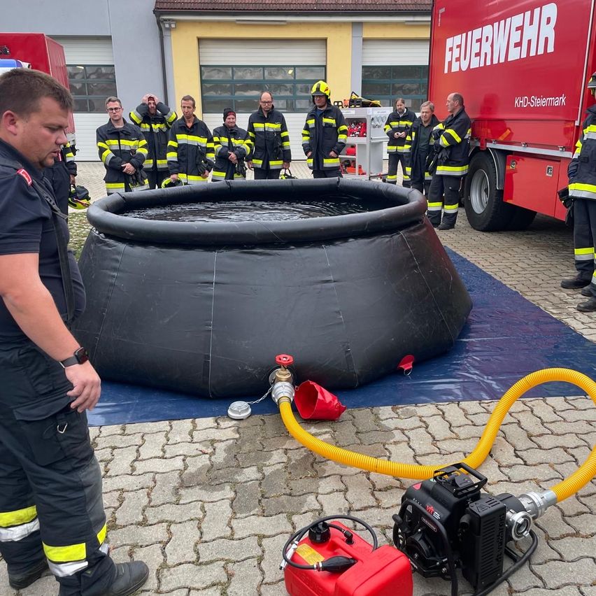 Eine Gruppe Feuerwehrleute steht um einen großen aufblasbaren Pool mit Wasser herum. Ein Mann mit einer Uhr steht neben einem roten Generator mit einem gelben Schlauch.