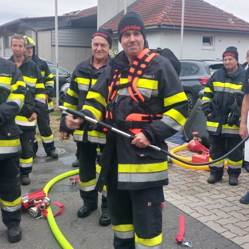 Mehrere Feuerwehrleute stehen zusammen auf einer nassen Straße. Ein Mann hält einen Schlauch. Dahinter stehen Feuerlöscher, ein Haus und geparkte Autos.