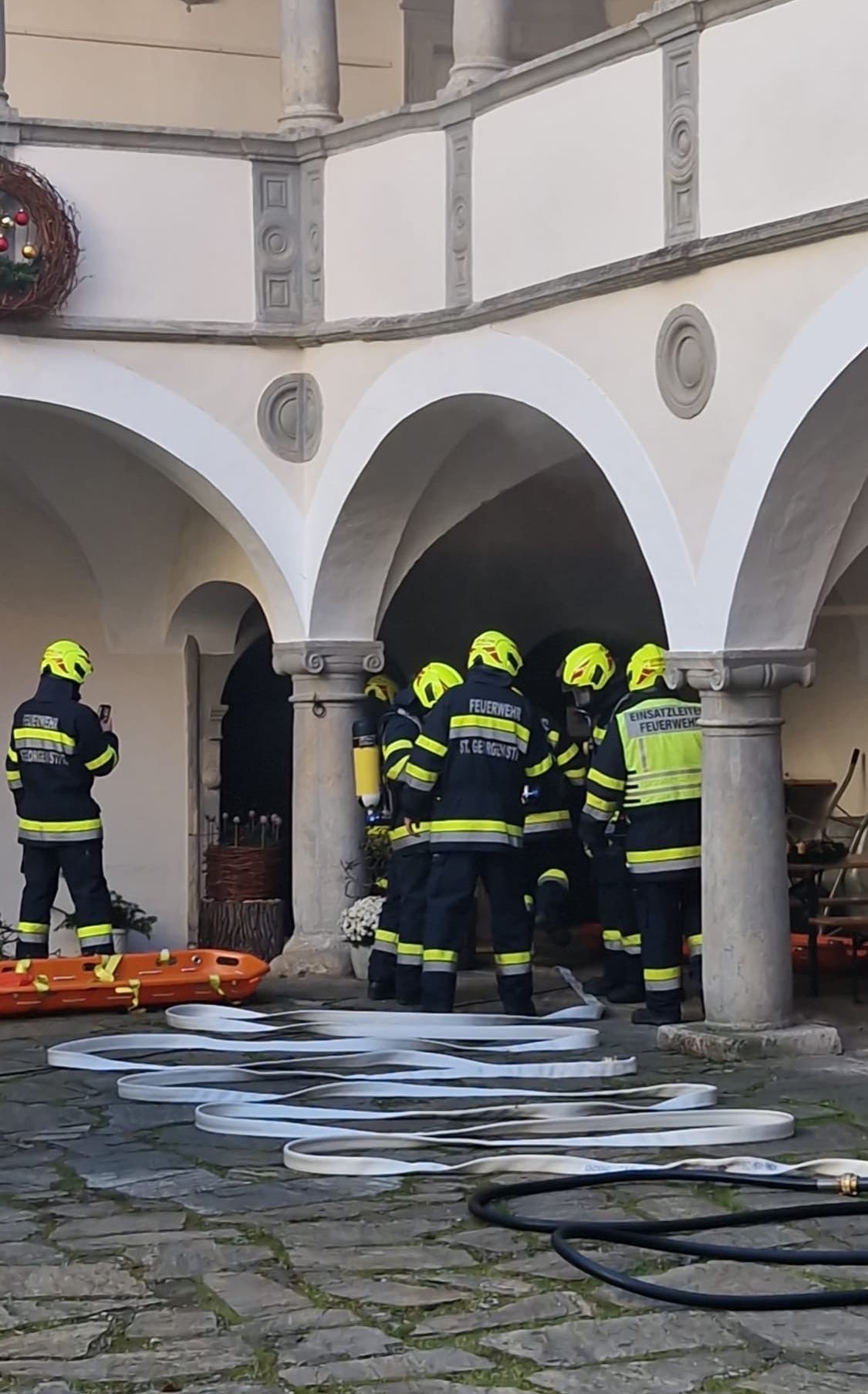 Feuerwehrleute in gelben Helmen und Uniformen stehen in einem Innenhof mit gebogenen Säulen. Ein Feuerwehrmann steht in der Nähe einer orangefarbenen Trage. Die Szene spielt sich in einem Gebäude mit gewölbten Eingängen ab.