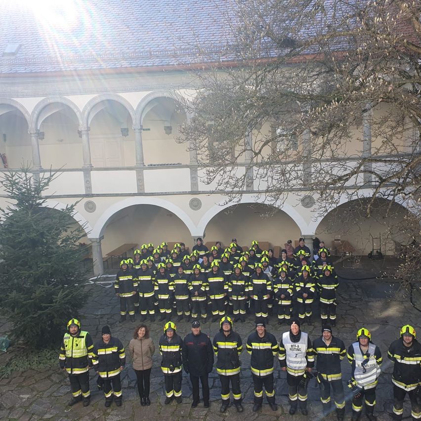Eine Gruppe Feuerwehrleute, gekleidet in gelben und schwarzen Uniformen, posiert für ein Foto vor einem historischen Gebäude. Links steht ein Weihnachtsbaum.