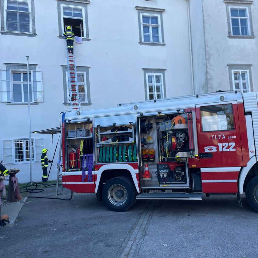 Ein Feuerwehrmann klettert eine Leiter zu einem Fenster eines Gebäudes. Ein Feuerwehrwagen mit der Nummer 122 ist in der Nähe geparkt.