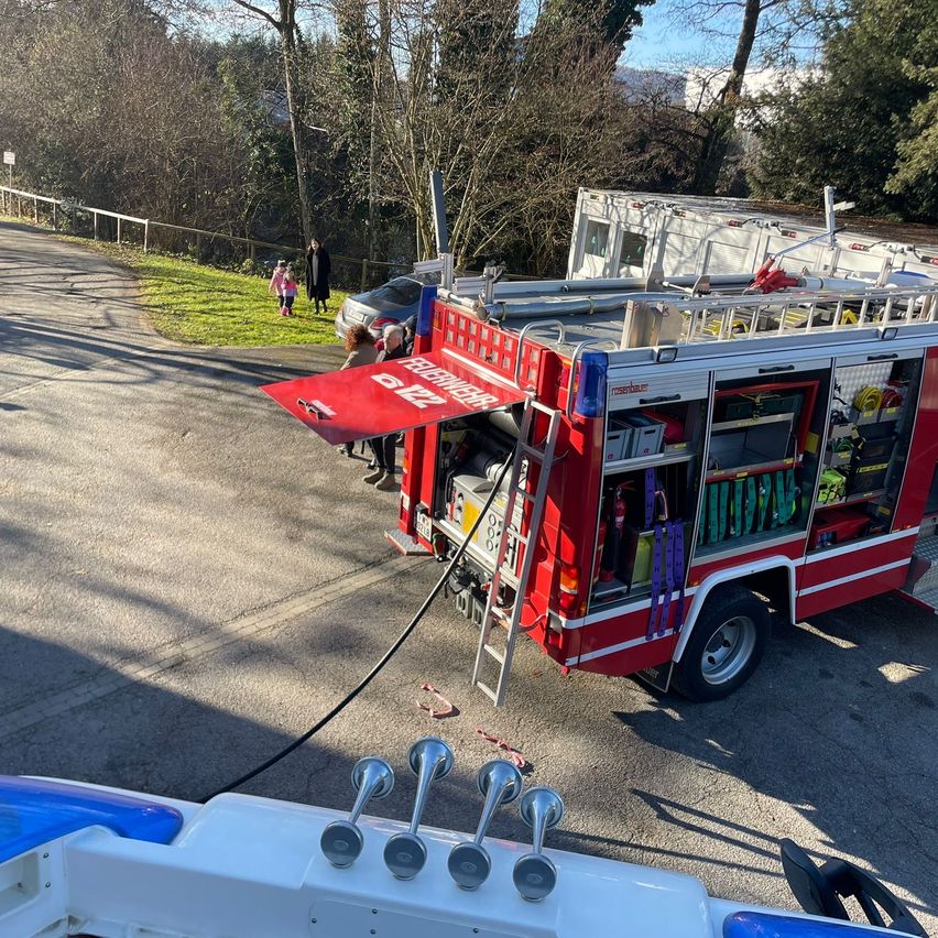 Ein roter Feuerwehrwagen mit Ausrüstung auf dem Dach steht am Straßenrand mit ausgefahrener Leiter. In der Nähe gehen Menschen auf dem Gras.
