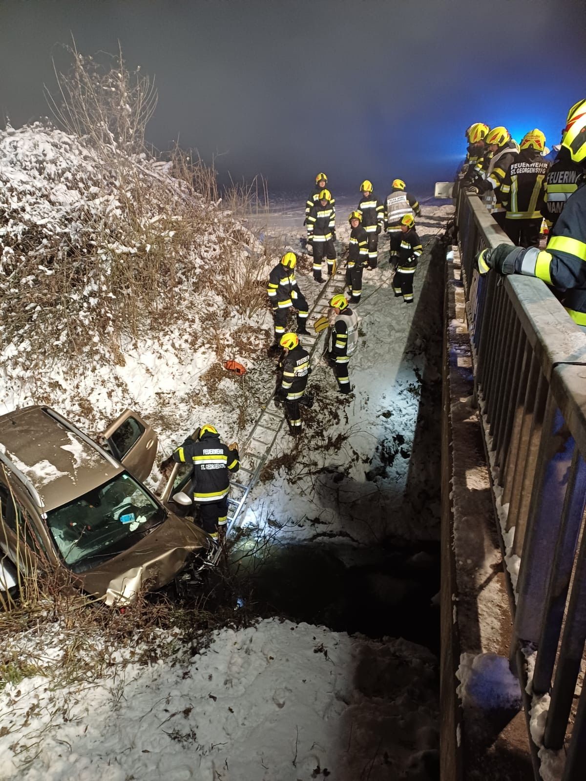 Ein Auto ist auf einer verschneiten Straße in einen Graben gestürzt, Feuerwehrleute in gelben und schwarzen Uniformen retten den Fahrer.