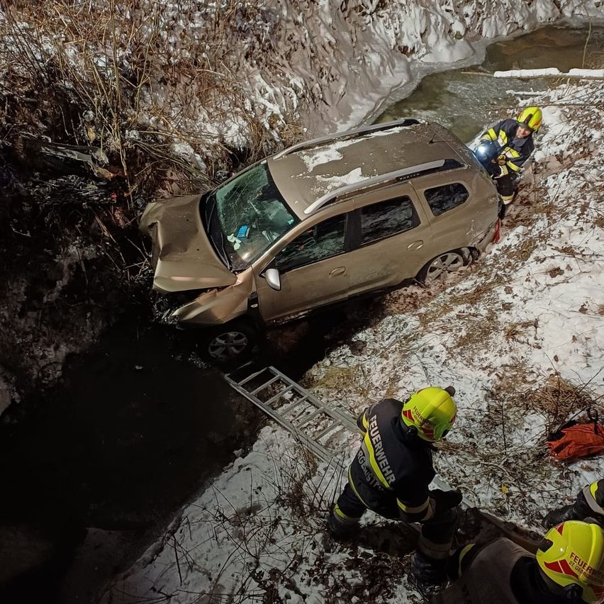Rettungskräfte retten ein Auto, das in einen Fluss gerutscht ist. Das Auto ist zur Hälfte im Wasser, und drei Personen tragen gelbe Helme.