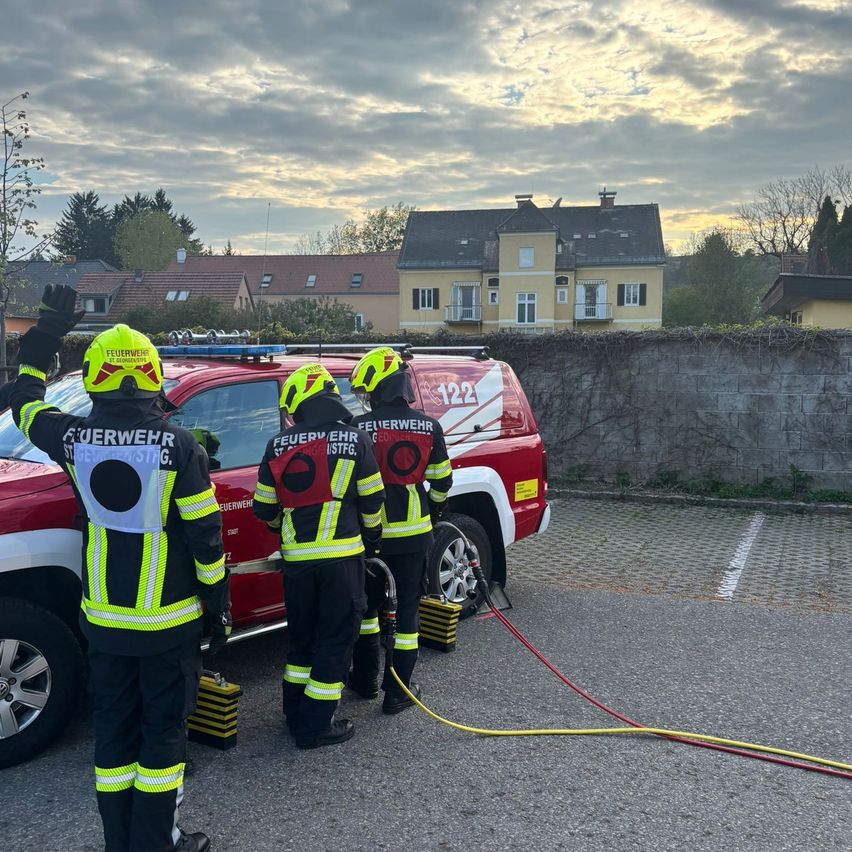 Three firefighters are preparing to handle a fire emergency. They stand by a red fire truck, with hoses and equipment in place. A wall with plants and a house are in the background.