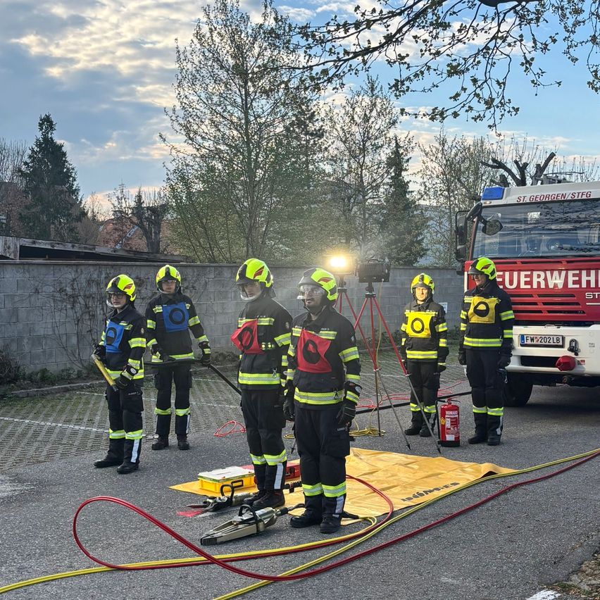 Firefighters in protective gear stand in a parking lot with a fire truck and a fire extinguisher nearby.