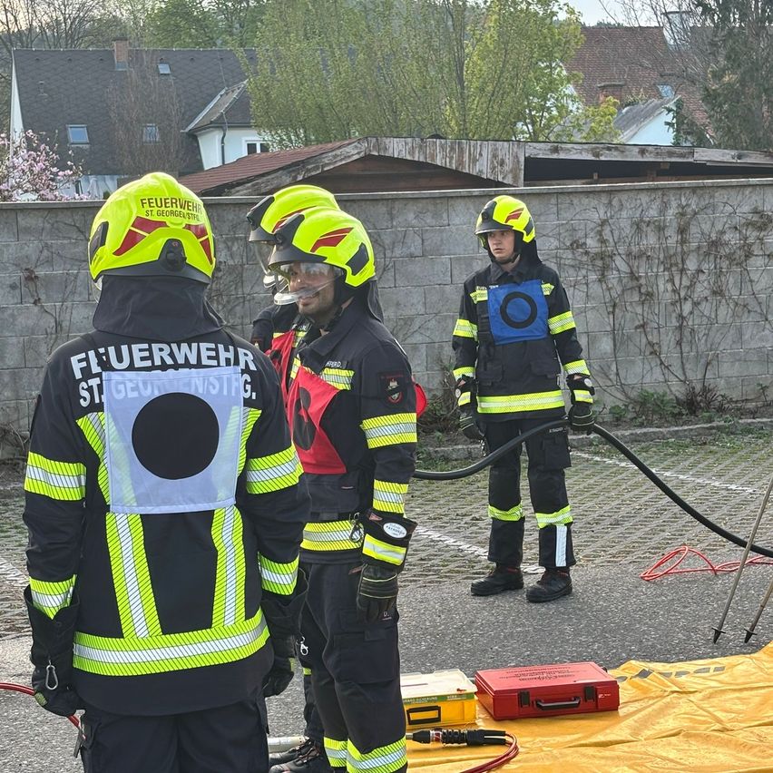 Three firefighters in full gear stand by a wall. One holds a hose while another observes. They are outside, with a yellow tarp and red box nearby.
