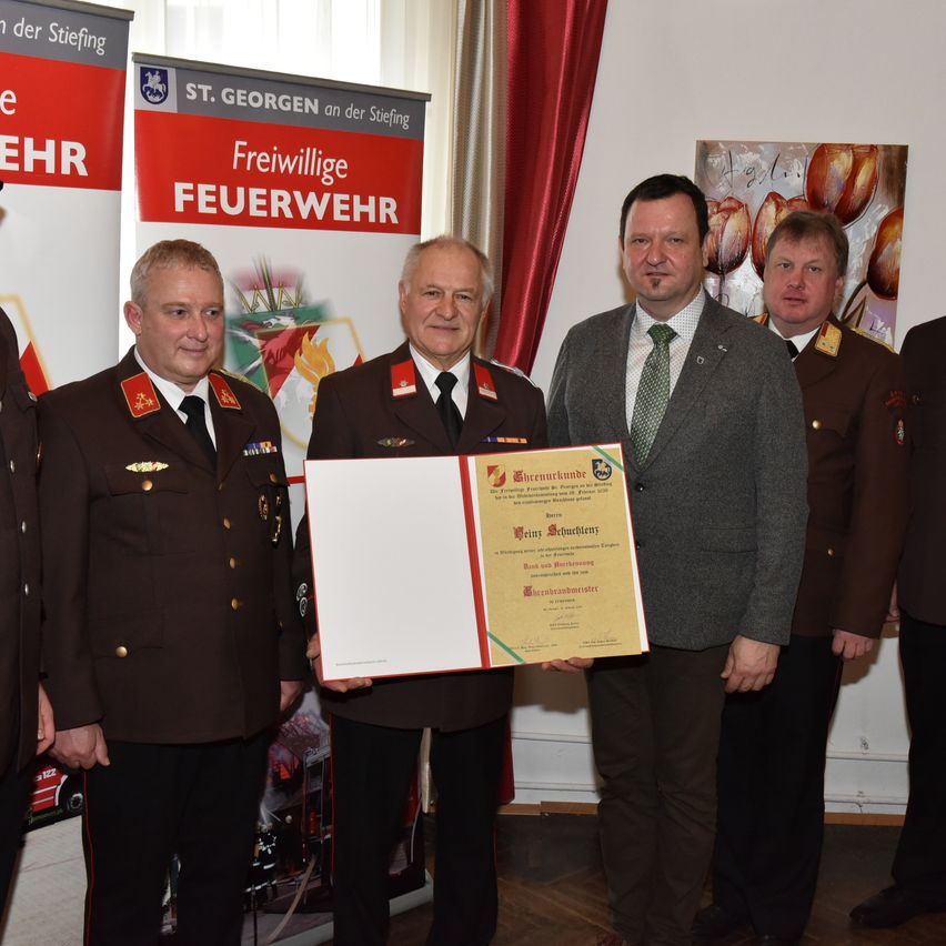 A man in a business suit presents a certificate to a man in uniform. Behind them, several people in uniforms stand, including one with a mustache. They are in front of a banner with the words 'Freiwillige Feuerwehr.'