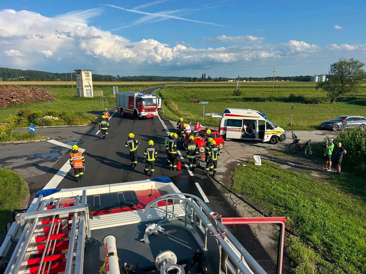 Rettungskräfte in gelben Helmen helfen bei einem Autounfall auf der Straße. Ein Krankenwagen ist geparkt, und ein Feuerwehrauto ist am Unfallort.