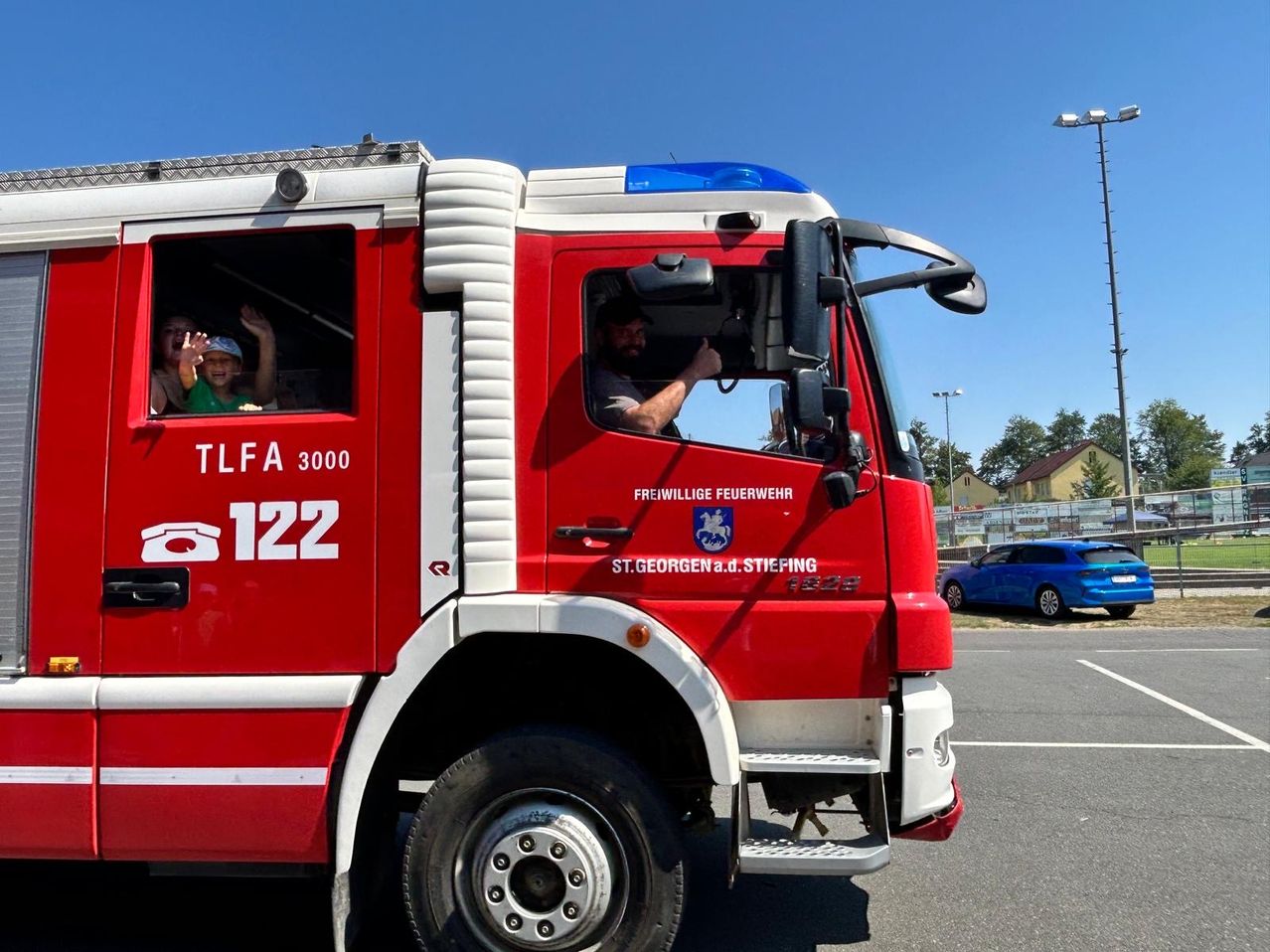 Ein roter Feuerwehrwagen mit der Nummer 122 vorne und an der Seite, mit einem Mann am Steuer und zwei Kindern hinten, die aus dem Fenster schauen.