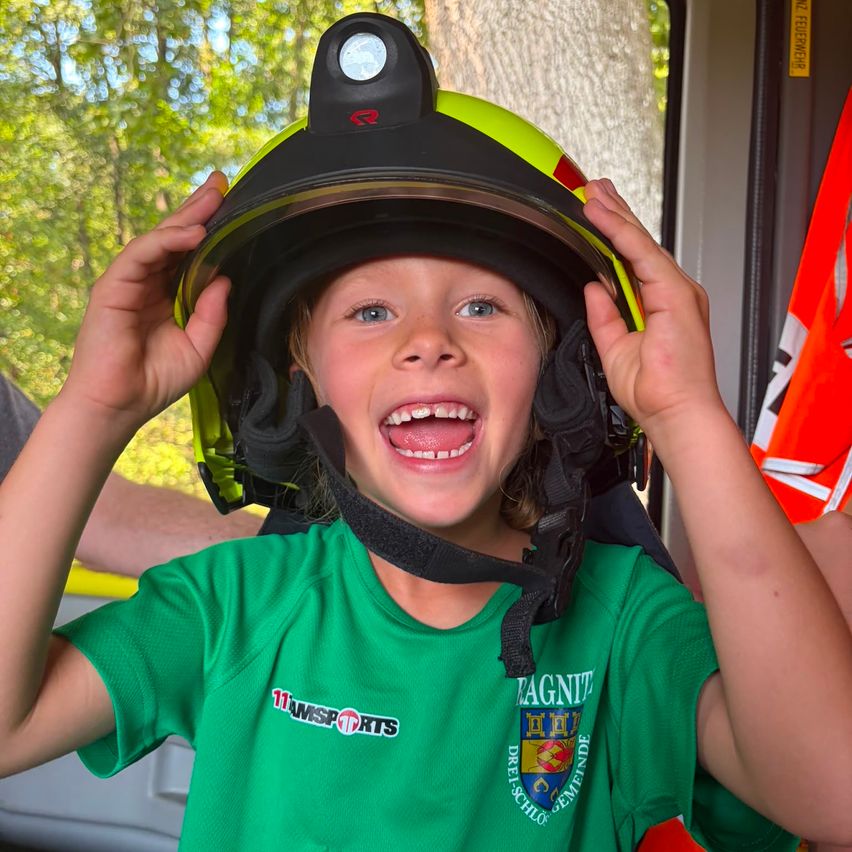 Ein junger Junge mit grünem Shirt und Feuerwehrhelm lächelt und hält den Helm mit beiden Händen.