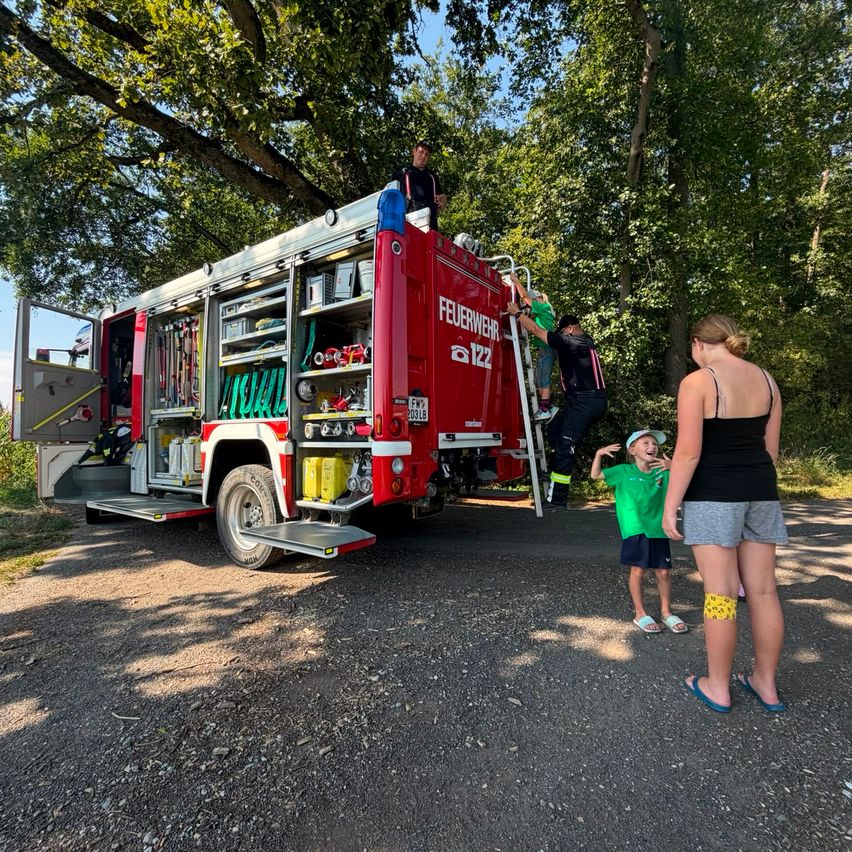 Ein roter Feuerwehrwagen mit offenen Türen ist auf einer Kiesstraße geparkt. Zwei Feuerwehrleute klettern auf einer Leiter an der Seite des Trucks, während ein Mann oben steht. Eine Frau und zwei Kinder stehen in der Nähe und beobachten.