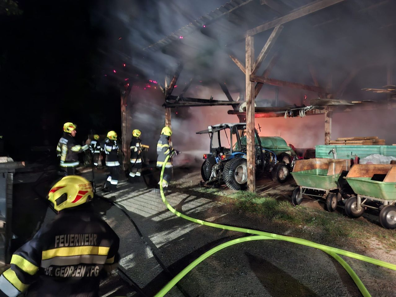 Feuerwehrleute in gelben Helmen bekämpfen ein Scheunenfeuer bei Nacht, mit Rauch, der aus dem Dach aufsteigt und einem Traktor in der Nähe.