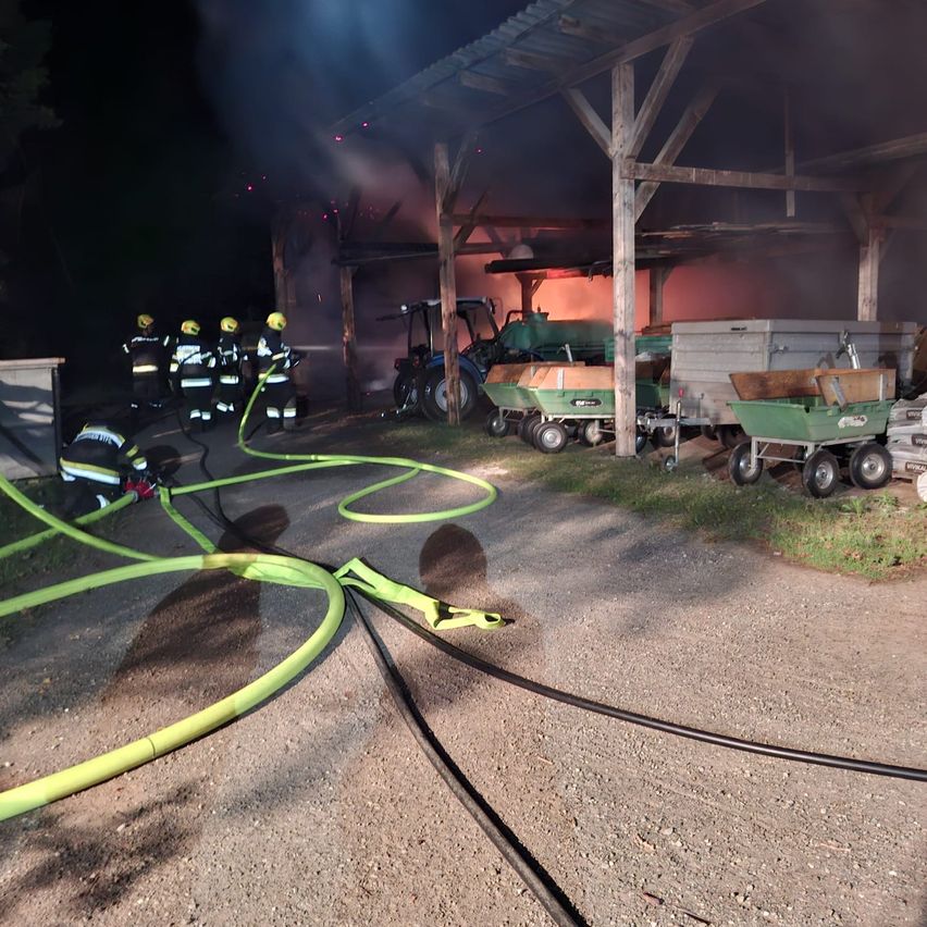 Feuerwehrleute mit gelben Helmen bekämpfen einen Scheunenbrand bei Nacht. Geräte und Fahrzeuge sind um die brennende Struktur herum geparkt.