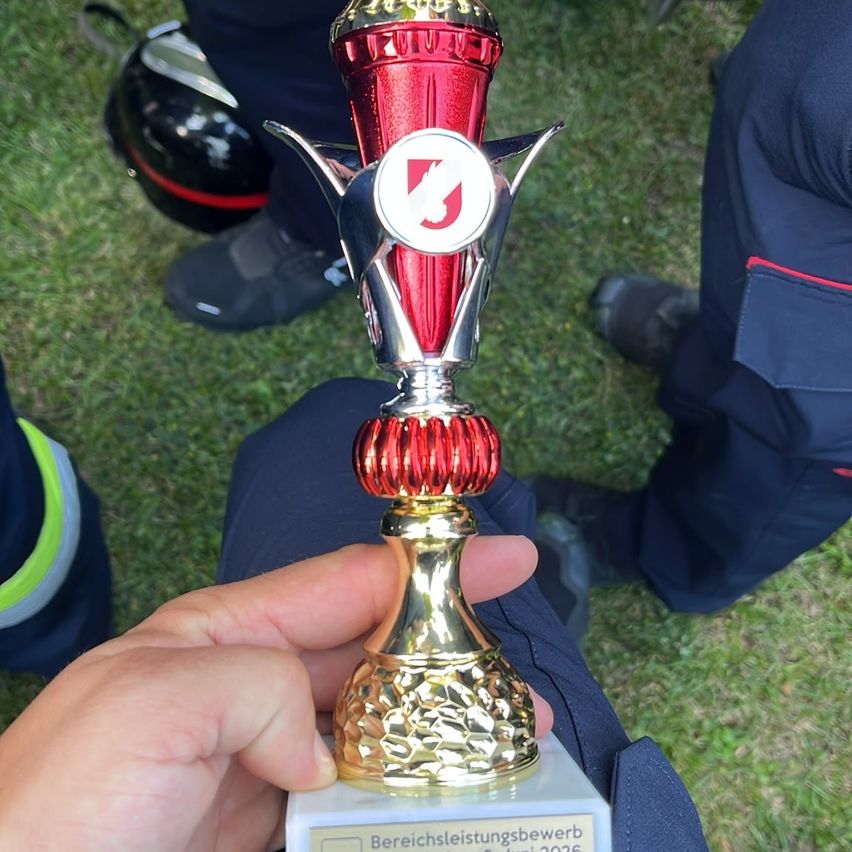 A person holds a trophy, red and gold, on a lawn. Two individuals in uniform sit behind the trophy holder.