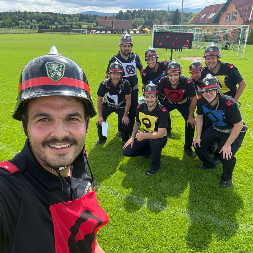A group of people in helmets and black and yellow shirts pose for a photo on a green field. They have numbers on their backs. Behind them is a soccer field and a scoreboard.