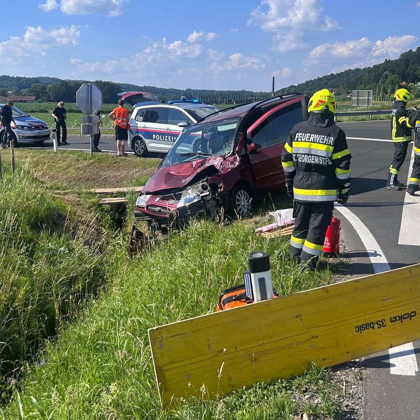 Rettungskräfte kümmern sich um ein auf der Straßenseite verunfalltes rotes Auto. Feuerwehrleute stehen neben dem Fahrzeug.