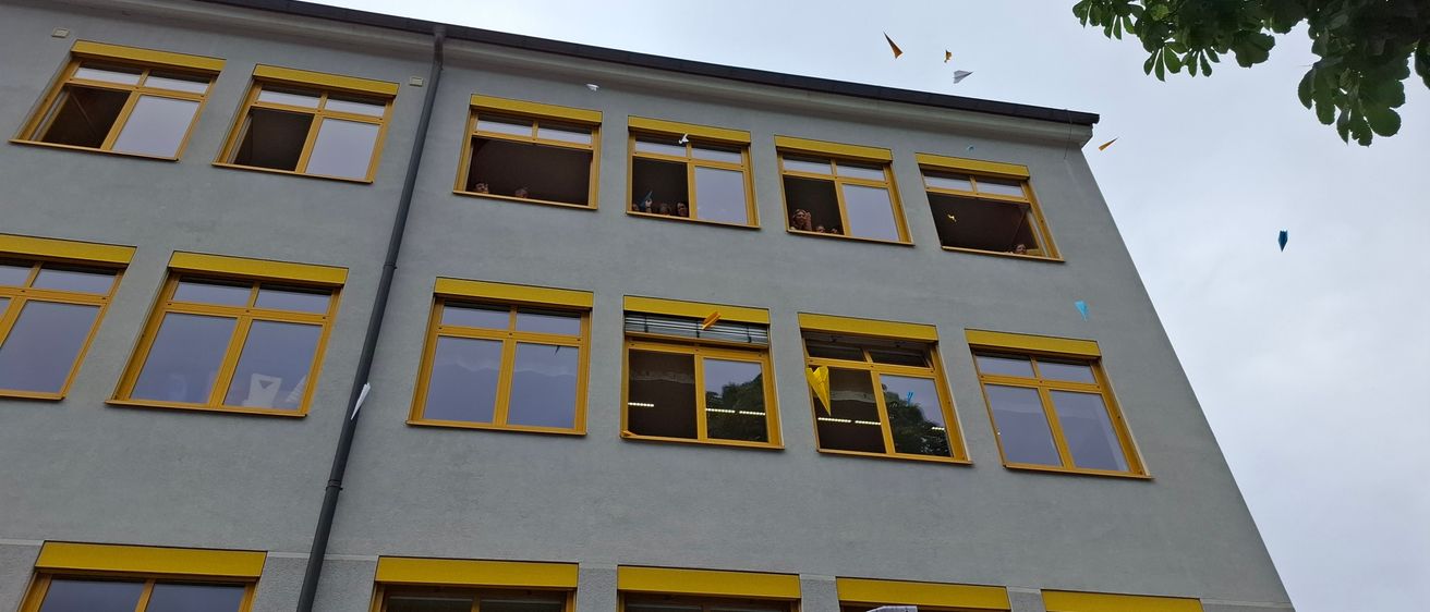 Ein mehrstöckiges Gebäude mit gelben Fensterrahmen und mehreren Fenstern. Einige Menschen schauen aus den Fenstern.