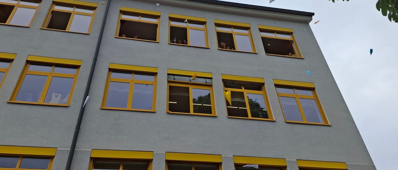Ein mehrstöckiges Gebäude mit gelben Fensterrahmen und mehreren Fenstern. Einige Menschen schauen aus den Fenstern.