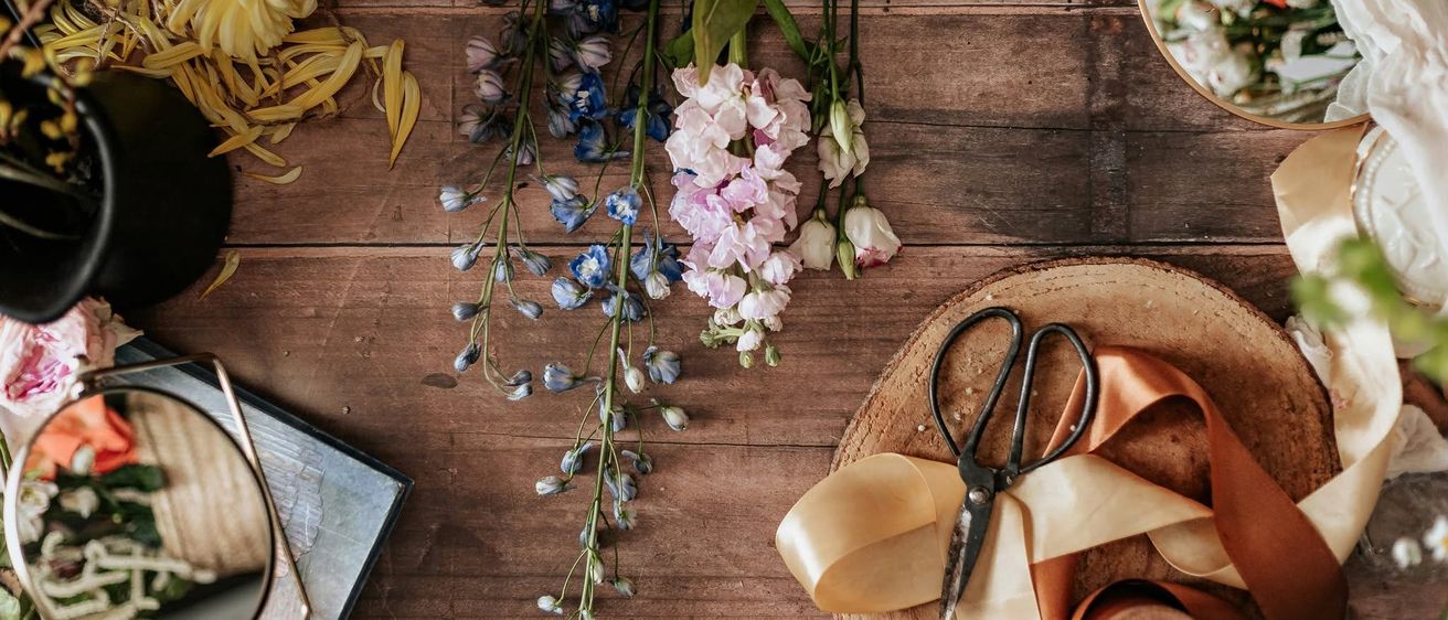 Ein Holztisch zeigt verschiedene Blumen und Stängel, Scheren und Bänder und deutet auf eine Bastel- oder Blumenarrangement-Einrichtung hin.