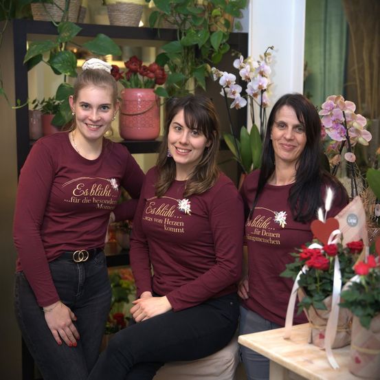 Drei Frauen in passenden weinroten Shirts posieren für ein Foto in einem Blumenladen, mit verschiedenen Pflanzen und Blumen im Hintergrund.