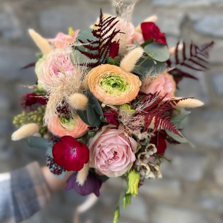 Ein Nahaufnahme einer Hochzeitsbouquet mit einer Mischung aus rosa, orange und roten Blumen, ergänzt durch grüne Blätter und getrocknete Elemente.