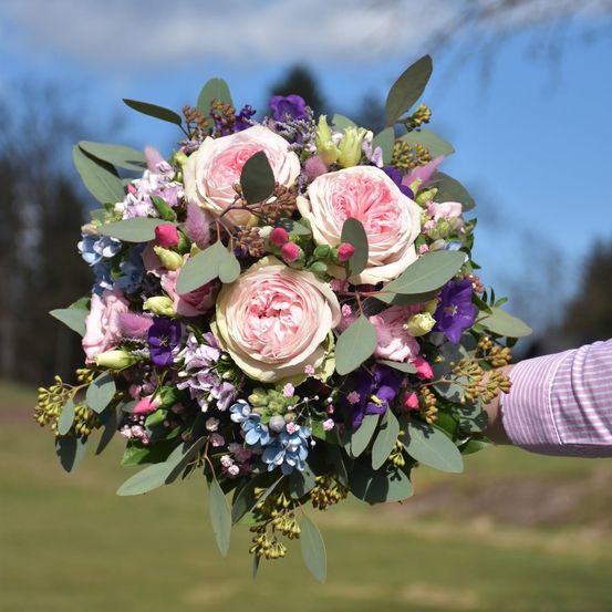 Ein Nahaufnahme eines farbenfrohen Blumenstraußes mit rosa Rosen, blauen Hortensien und grünen Blättern, gehalten von einer Person auf einer Wiese unter einem blauen Himmel.