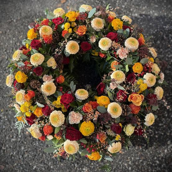 Ein kreisförmiger Kranz aus verschiedenen Blumen wie Rosen, Gänseblümchen und anderen saisonalen Blüten, auf einer Kiesoberfläche angeordnet.