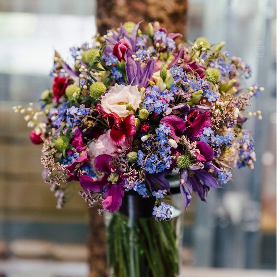 Ein farbenfroher Strauß aus verschiedenen Blumen, darunter lila, rosa und blaue Blüten, präsentiert in einem klaren Glasvase.
