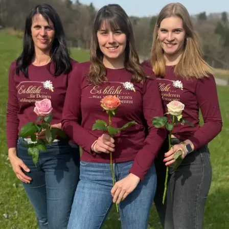 Drei Frauen in passenden weinroten Shirts posieren für ein Foto auf einem Feld, jede hält eine Rose in der Hand. Die Frau in der Mitte hat eine Rose in der Hand und ein Lächeln im Gesicht.