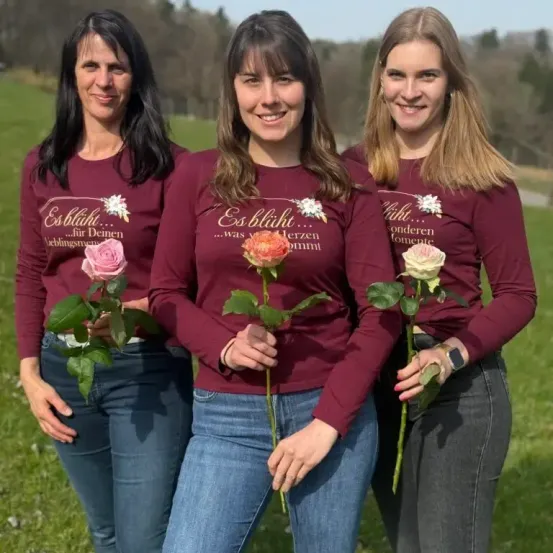 Drei Frauen in passenden weinroten Shirts posieren für ein Foto auf einem Feld, jede hält eine Rose in der Hand. Die Frau in der Mitte hat eine Rose in der Hand und ein Lächeln im Gesicht.