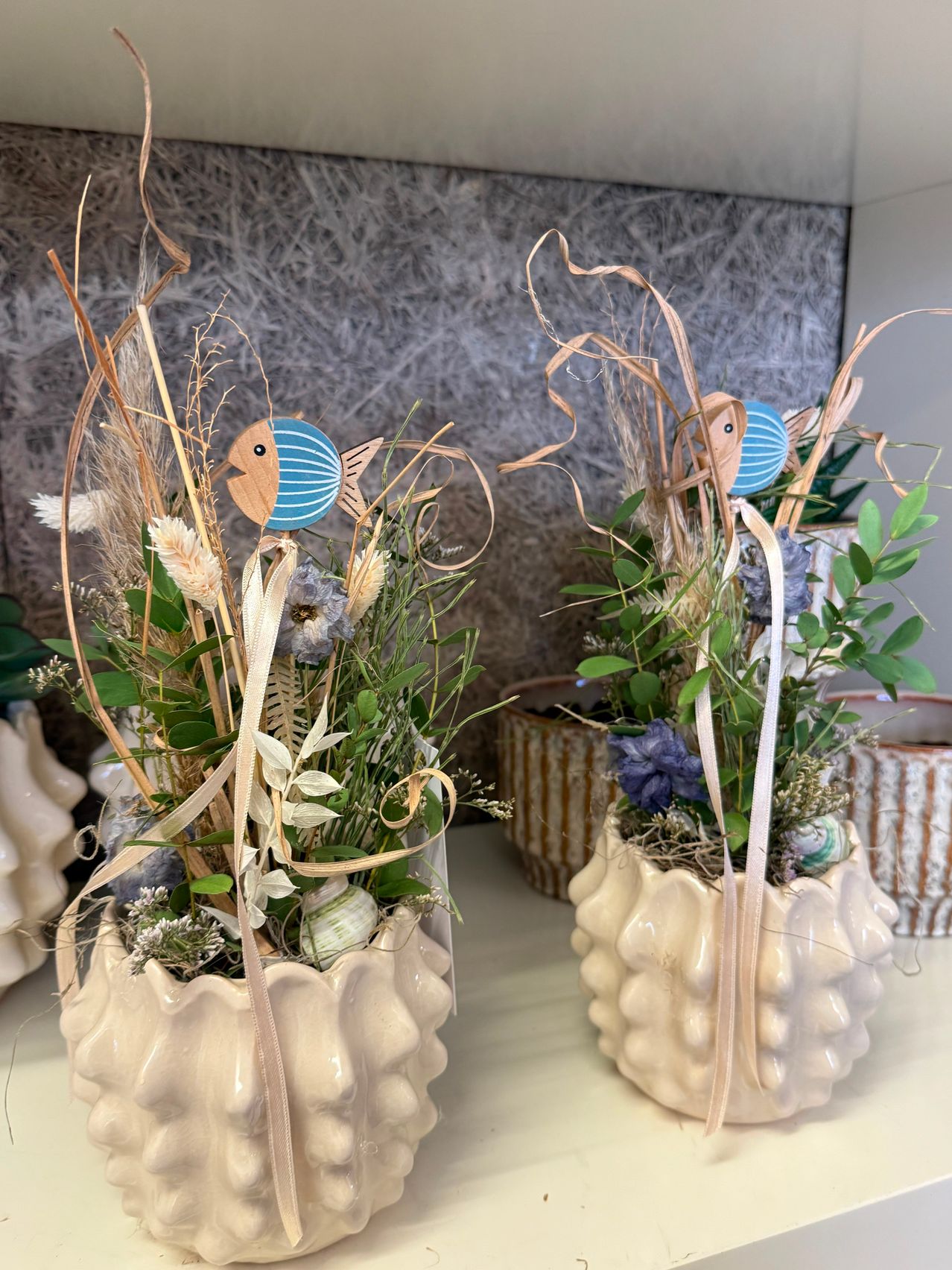 Zwei dekorative Keramikvasen mit Blumenarrangements und blauen Fischfiguren oben, auf einer weißen Oberfläche ausgestellt.