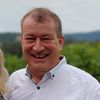 Ein Mann und eine Frau stehen in einem Weinberg, lächeln und halten Weingläser. Der Mann trägt ein weißes Hemd, und die Frau trägt eine Jeansjacke. Sie stehen vor einem Hintergrund aus üppigem Grün und einem bewölkten Himmel.