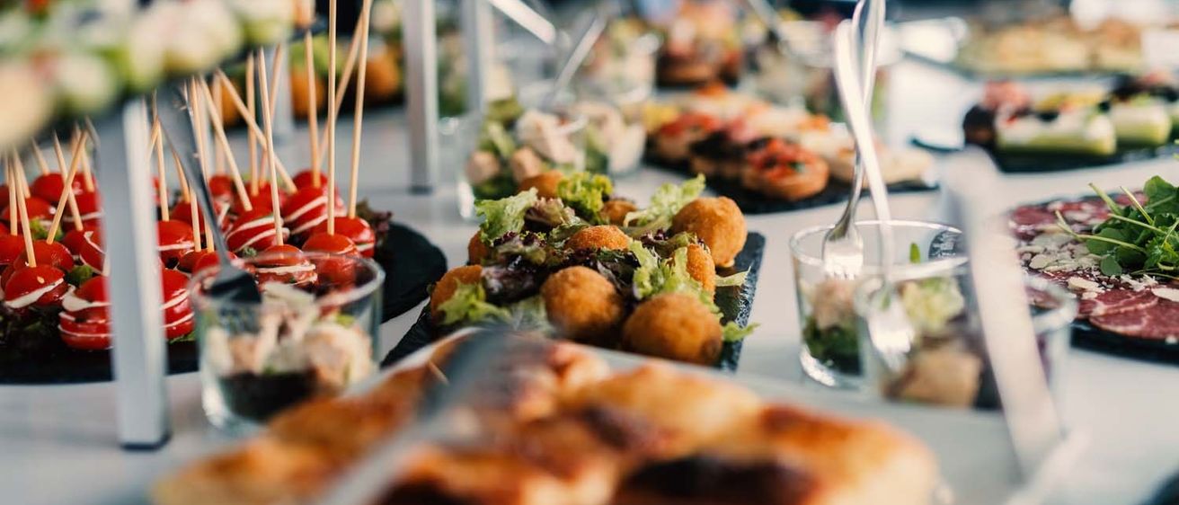 Ein Buffettisch zeigt eine Vielzahl von Vorspeisen. Es gibt Mini-Sandwiches, einen Salat mit Salat und Hähnchenbällchen und Tomaten-am-Stielen-Spieße.