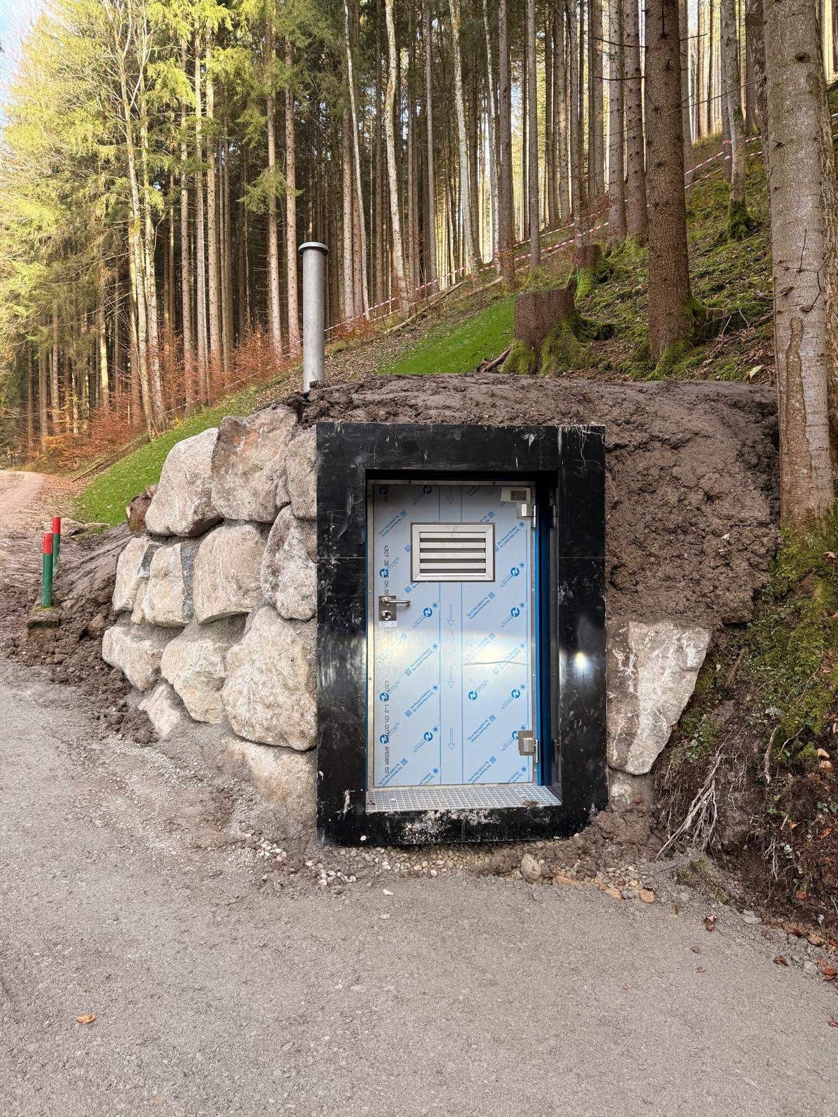 Eine kleine Tür ist in eine Steinmauer eingebettet. Sie hat einen grauen Metallrahmen und ist von Erde umgeben. Ein Metallrohr ist auf der Erde platziert. Die Mauer besteht aus Steinen und befindet sich auf einem geneigten Bereich mit einem Wald.