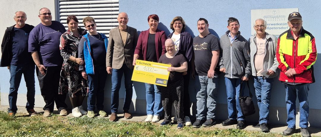 A group of smiling adults stand behind a person holding a large check. The person holds a yellow check with the text 'x Scheck'. They are all wearing casual clothing.